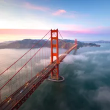 An aerial view of the Golden Gate Bridge with Karl the fog running under it and a beautiful sky with hues of pink yellow and blue in the background