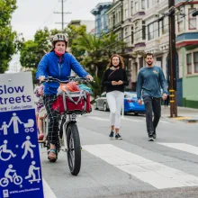 A mother pedals a bicycle with two children riding behind her, while two pedestrians walk behind, all in front of an SFMTA Slow Streets sign