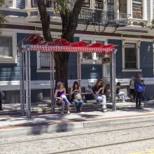 People waiting under a red awning on a bench at a bus stop