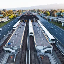 BART trains at a center platform station between freeway lanes in the San Francisco Bay Area, with cars on either side and hills in the background at sunset.