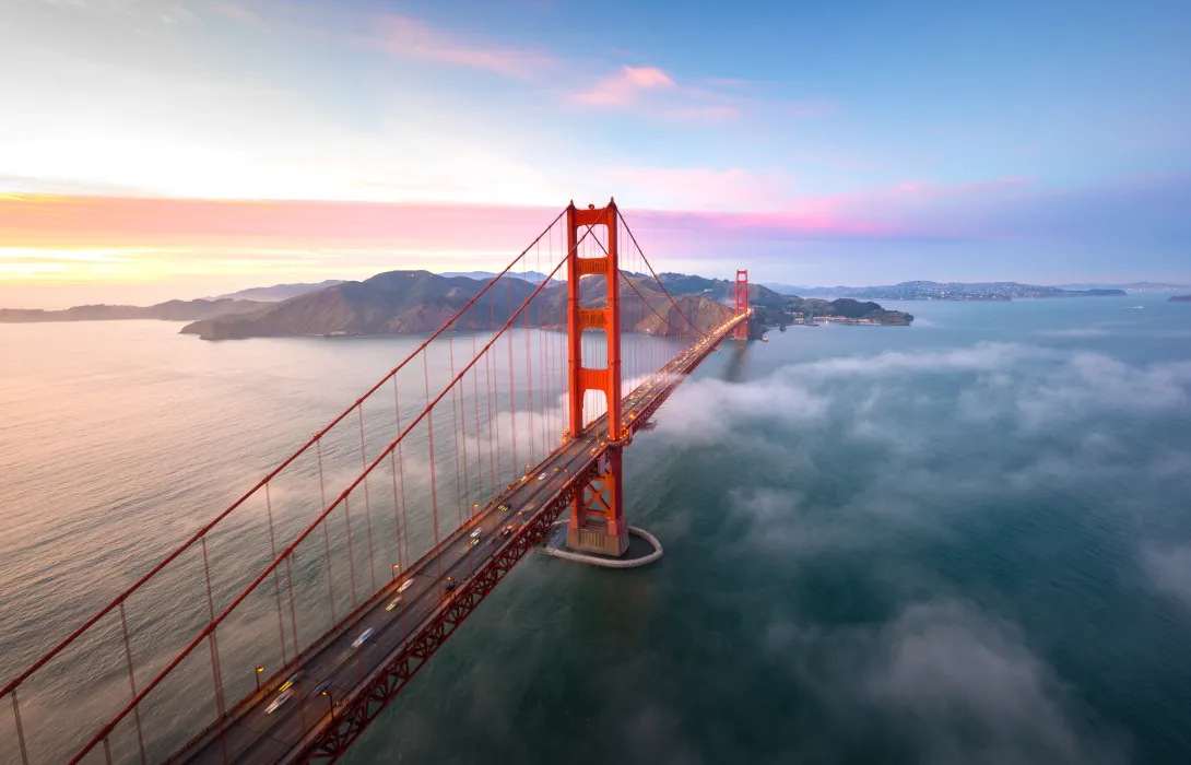 An aerial view of the Golden Gate Bridge with Karl the fog running under it and a beautiful sky with hues of pink yellow and blue in the background