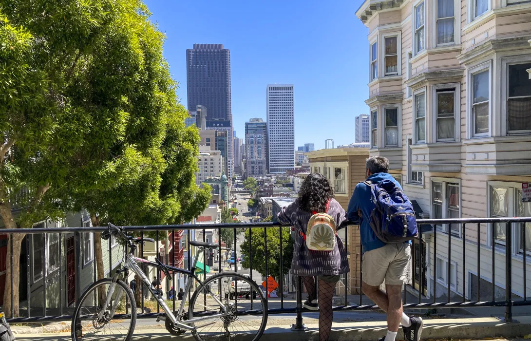 Two people next to a bicycle peer down over a railing at a bustling cityscape below