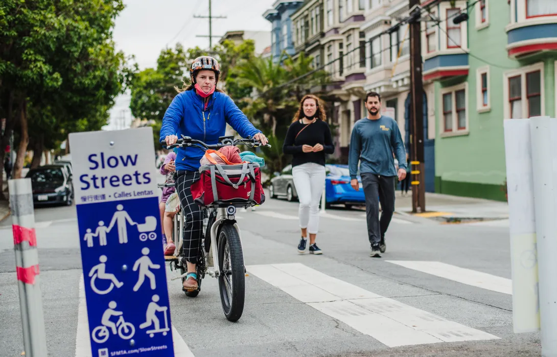 A mother pedals a bicycle with two children riding behind her, while two pedestrians walk behind, all in front of an SFMTA Slow Streets sign