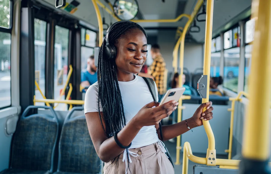 A smiling woman wearing headphones grips a bus handrail and smiles at her smartphone as she rides the bus