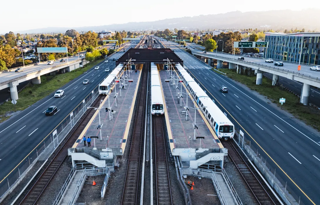 BART trains at a center platform station between freeway lanes in the San Francisco Bay Area, with cars on either side and hills in the background at sunset.